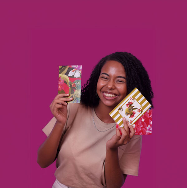Mulher jovem negra com cabelos curtos sorri ao exibir envelopes e cartões estampados com estampas florais e da fauna. O fundo rosa-violeta vibrante realça a cena. 