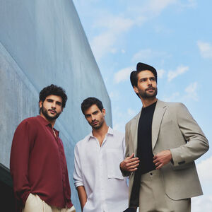 Três homens jovens de cabelos e barbas castanhos, à esquerda de camisa vinho, de camisa branca no centro e à direita de terno cáqui e camisa preta, em fundo de concreto e céu azul.