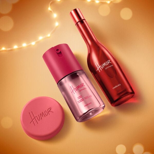 Frasco de perfume, sabonete e desodorante Natura Humor sobre fundo amarelo com luzes pisca-pisca. As embalagens são em tons vibrantes de rosa e vermelho.