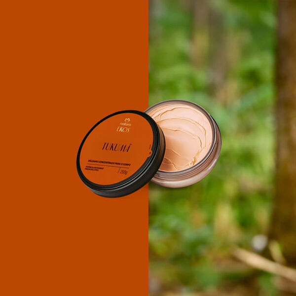 Pote aberto do Bálsamo Concentrado para o Corpo Ekos Tukumã, tampa laranja e creme bege denso. Fundo dividido entre laranja sólido e floresta desfocada ao fundo.