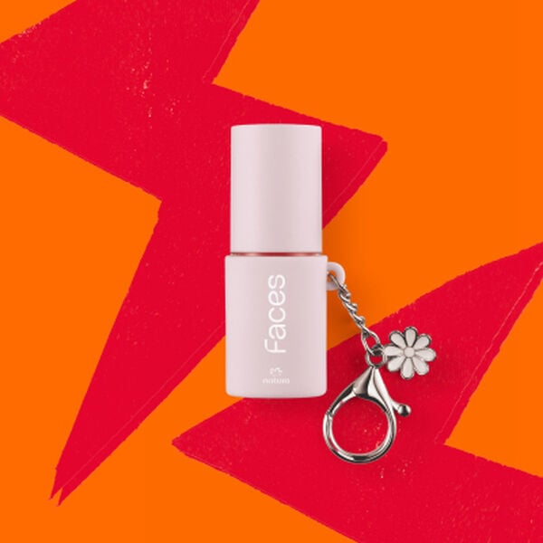Batom Natura Faces em embalagem rosa-claro acoplado a um chaveiro metálico com pingente de flor. O item está centralizado sobre um fundo vibrante com formas geométricas em laranja e vermelho.