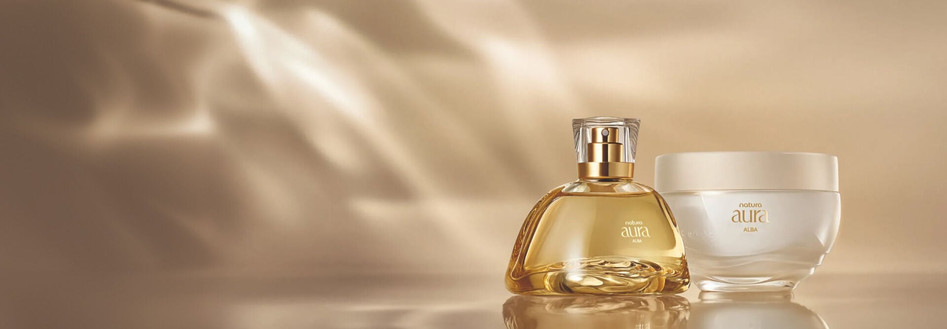 Frasco de perfume com líquido dourado e pote de creme hidratante da linha Natura Aura Alba sobre uma superfície clara e espelhada. Fundo em tom bege e acetinado com uma iluminação suave e sofisticada.
