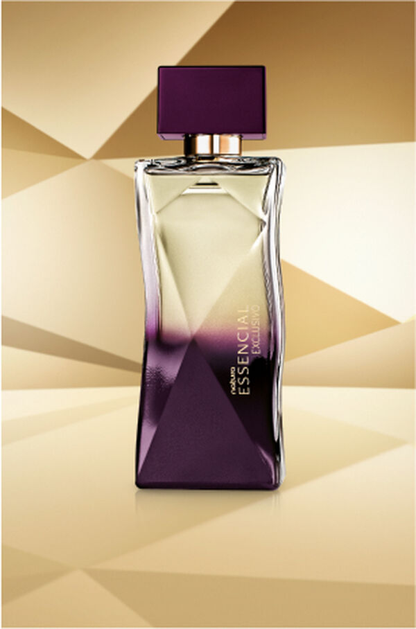 Frasco de Essencial Exclusivo Feminino, com topo transparente e base roxa, aparece em destaque no centro da tela sobre fundo multifacetado dourado, fazendo alusão ao design do frasco de Essencial.