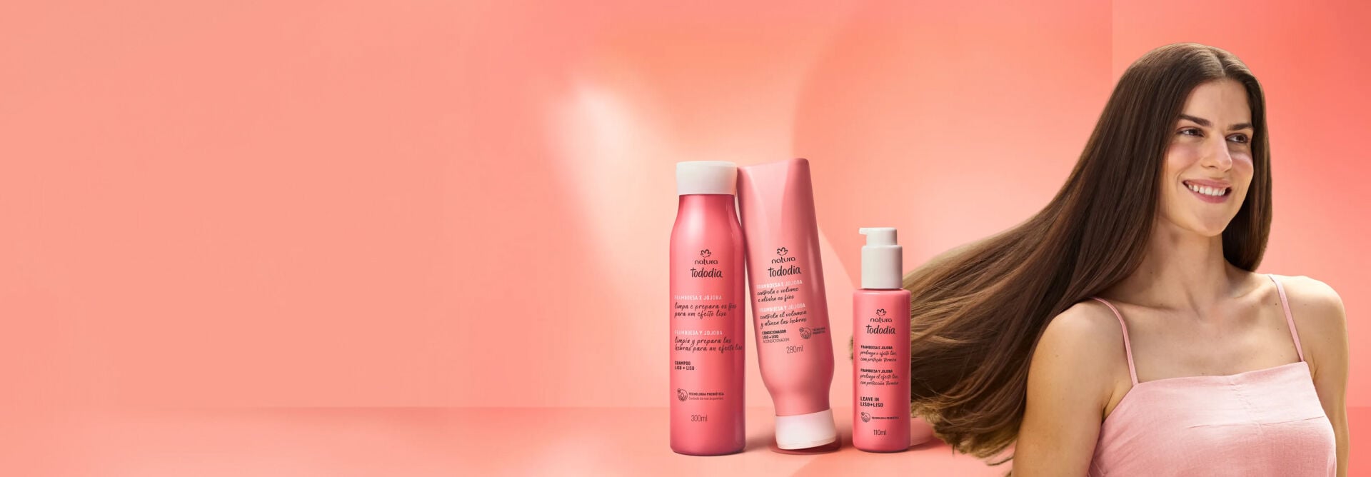 Mulher de pele clara sorri exibindo o movimento dos cabelos lisos, longos e castanhos. Ao lado, a linha Natura Tododia Framboesa e Jojoba: shampoo, condicionador e leave-in sobre fundo rosa.