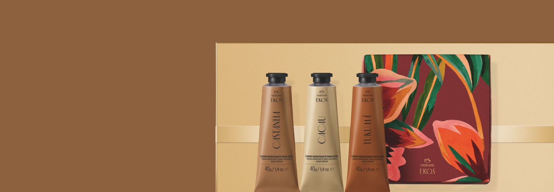 Três bisnagas de creme para mãos Natura Ekos (Castanha, Cacau e Tukumã) estão lado a lado. Ao fundo, uma caixa decorada com ilustrações florais sobre embalagem presenteável dourada.