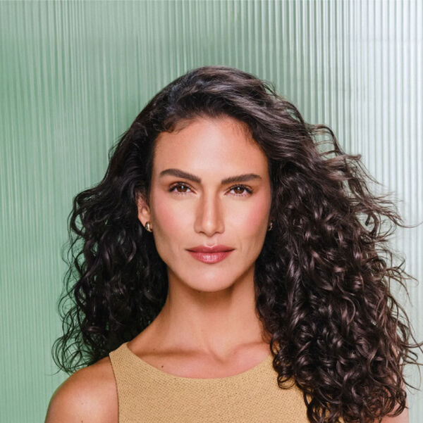 Mulher branca de cabelos soltos ondulados usa regata bege e segura produto Natura Lumina próximo ao corpo. Fundo em tons de verde com listras verticais.