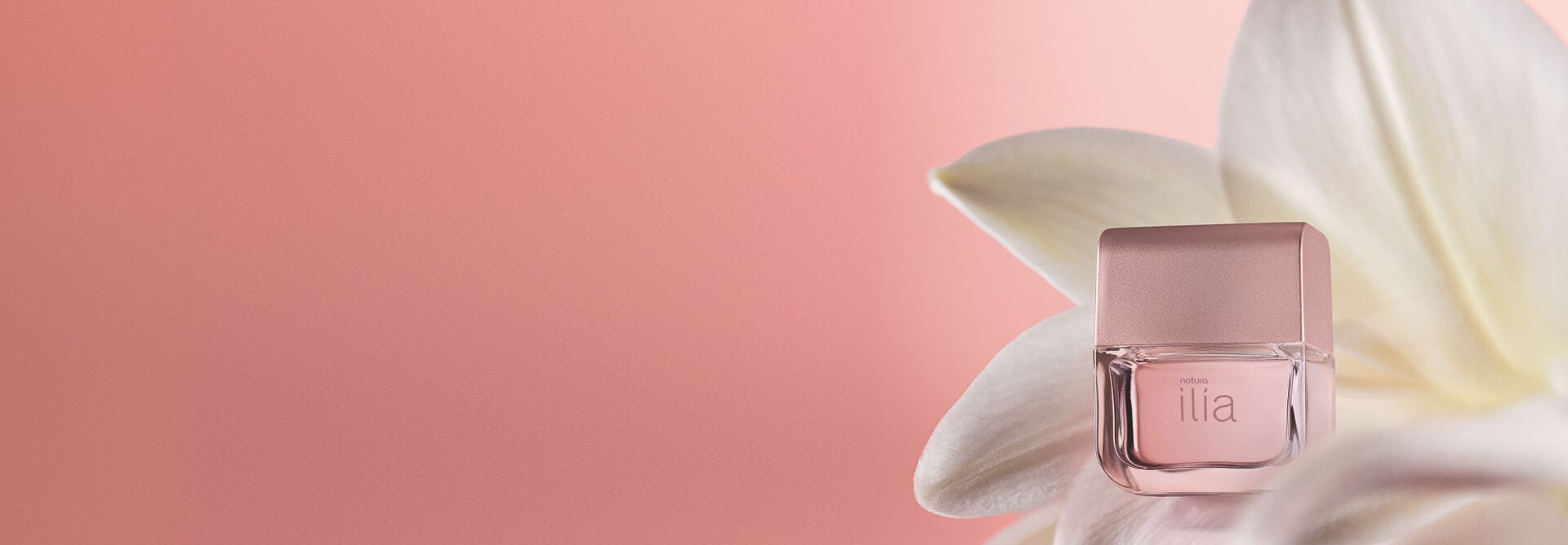 Frasco de perfume Natura Ilía repousa no centro de pétalas brancas de flor sobre fundo rosa. O vidro transparente e a tampa rosada destacam o tom delicado da fragrância.