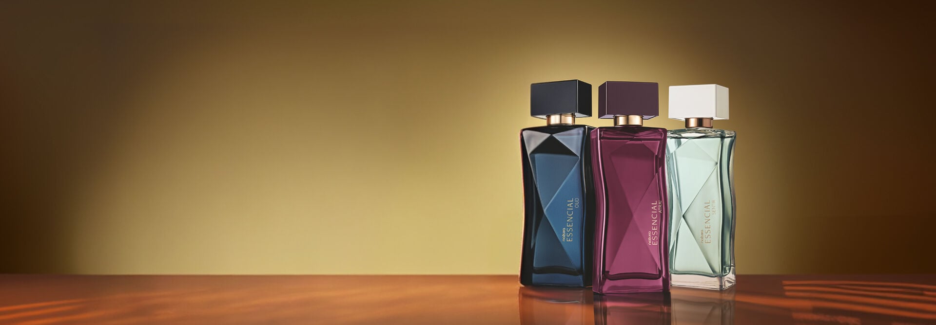 Três frascos de Natura Essencial estão alinhados sobre superfície espelhada em fundo dourado iluminado. As embalagens facetadas surgem nas cores azul-escuro, vinho e verde-claro.
