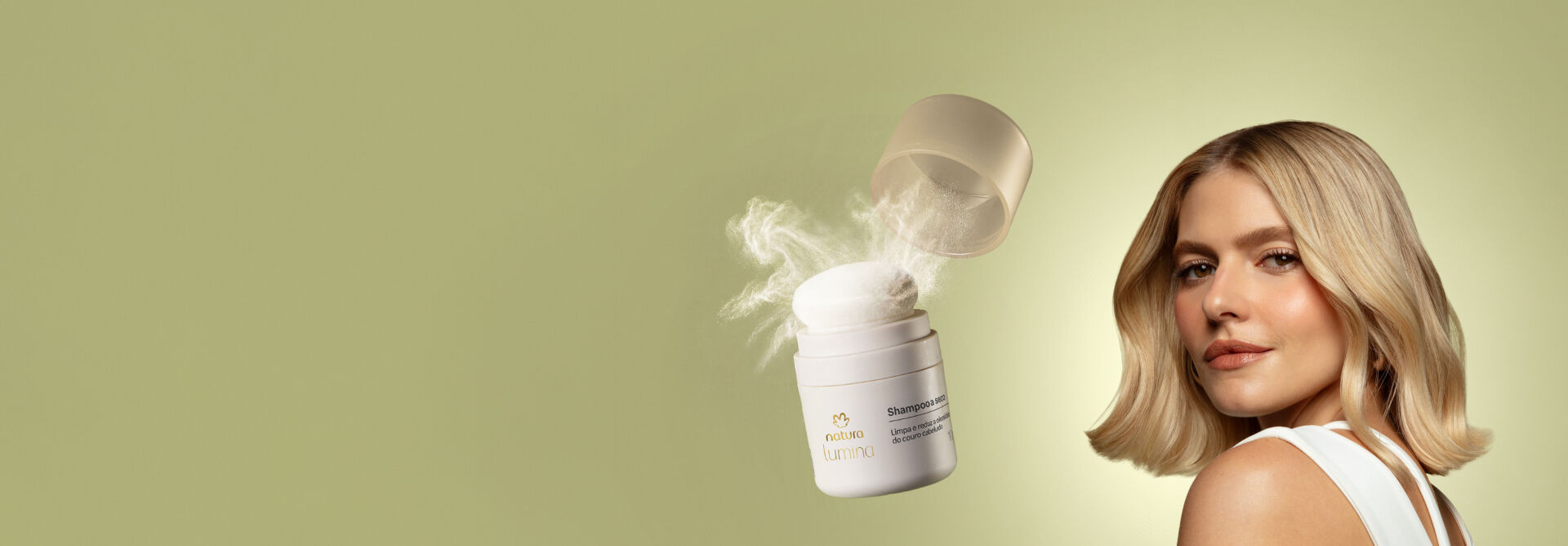 Mulher branca com cabelo loiro curto ondulado olha para a câmera em fundo verde-claro. Ao lado, frasco de Shampoo a Seco Natura Lumina aberto com aplicador de esponja liberando pó branco.