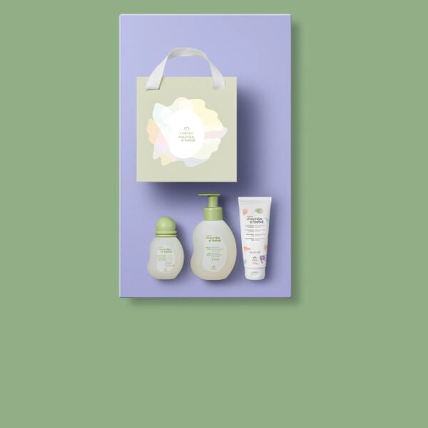Kit Natura Mamãe e Bebê organizado sobre fundo lilás. Acima, sacola de presente verde-clara; abaixo, colônia, sabonete líquido com pump e bisnaga de creme para assaduras alinhados.