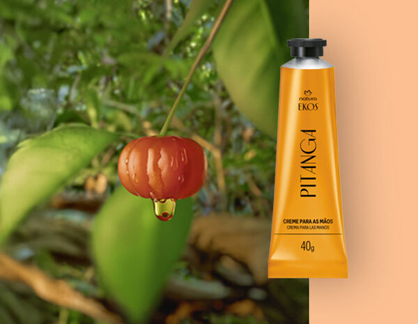 Bisnaga amarela do Creme Hidratante para as Mãos Ekos Pitanga à direita, ao lado de uma pitanga vermelha com gota de extrato, sobre fundo de folhas verdes e laranja suave.
