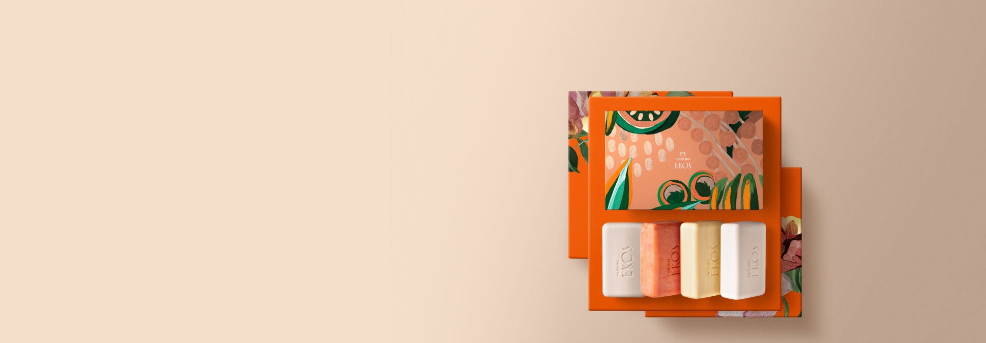 Presente Natura Ekos Sabonetes Sortidos, incluindo 1 castanha, 1 maracujá, 1 pitanga, 1 uccuba e 1 caixa presenteável decorada sobre caixas de presentes laranja com estampas de flores. Fundo bege. 