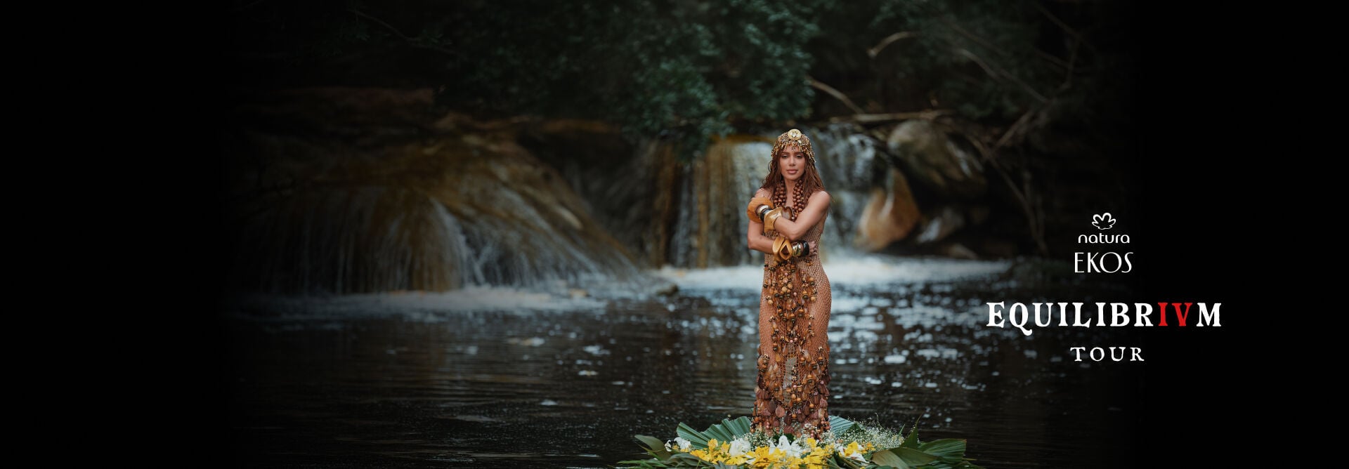 A cantora Anitta em uma cachoeira sobre oferenda floral, ao lado do logotipo Natura Ekos e do texto "Equilibrivm Tour".