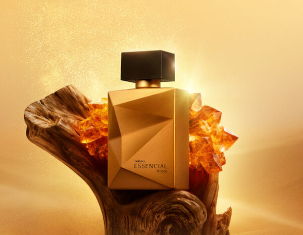 Deo Parfum Essencial Mirra Masculino em frasco facetado dourado com tampa preta. Está em fundo areia sobre um tronco de madeira escura e pedras âmbar ao redor.