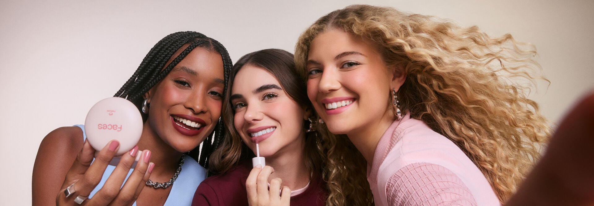 Três mulheres sorridentes posam juntas para uma foto estilo selfie. A mulher da esquerda, negra com tranças nagô, segura um pó compacto da linha Faces. A do meio aplica gloss nos lábios e a da direita, loira de cabelos cacheados, sorri olhando para a câmera. 