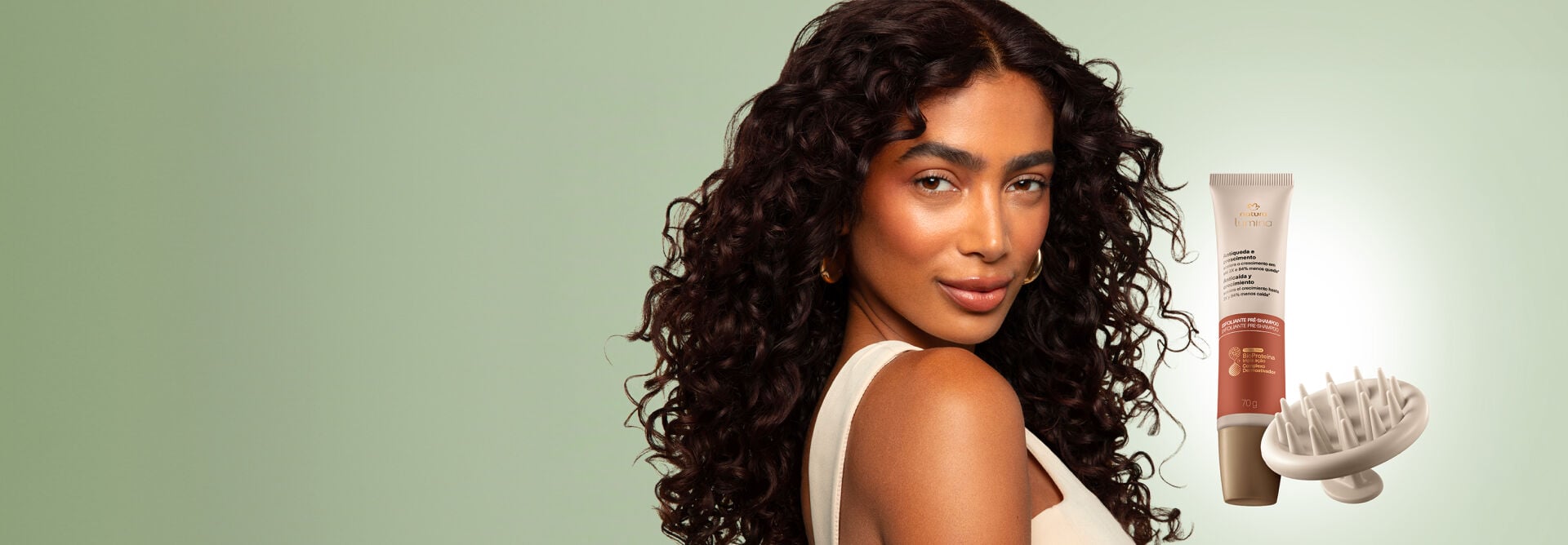 Mulher negra de cabelos cacheados volumosos olha para a frente em fundo verde. Ao lado, bisnaga do Esfoliante Pré-shampoo Natura Lumina Antiqueda e Crescimento com massageador capilar.