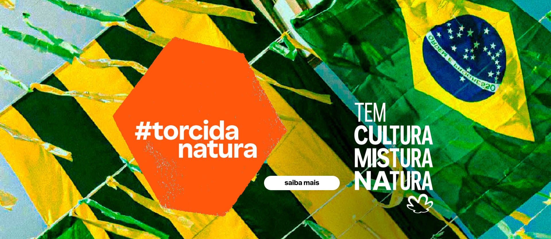 Fundo de céu azul com uma bandeira do Brasil e fitinhas verdes e amarelas. Hexágono laranja com o texto: tem Brasil na rua. Texto aplicado ao fundo: tem cultura mistura Natura.
