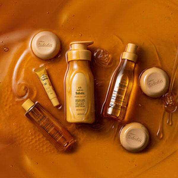 Produtos Natura Tododia Néctar de Mel imersos em mel líquido amarelo âmbar. A linha inclui hidratante, body splash, óleo corporal, sabonetes em barra e gloss labial.