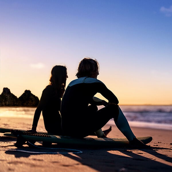 Silhueta de duas pessoas sentadas em pranchas de surfe na areia de uma praia durante o pôr do sol. Elas estão de costas, observando o mar sob um céu com tons de azul, dourado e laranja.