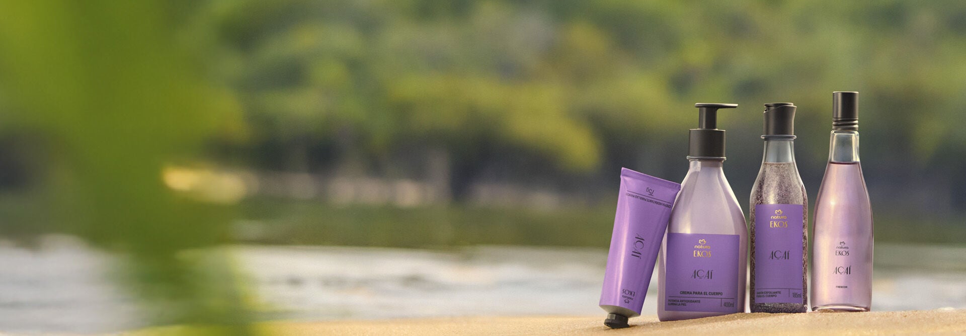 Quatro produtos Natura Ekos Açaí em embalagens lilás estão sobre a areia, com bisnaga, frasco pump, esfoliante e frasco de vidro, em frente a rio e floresta ao fundo desfocado.