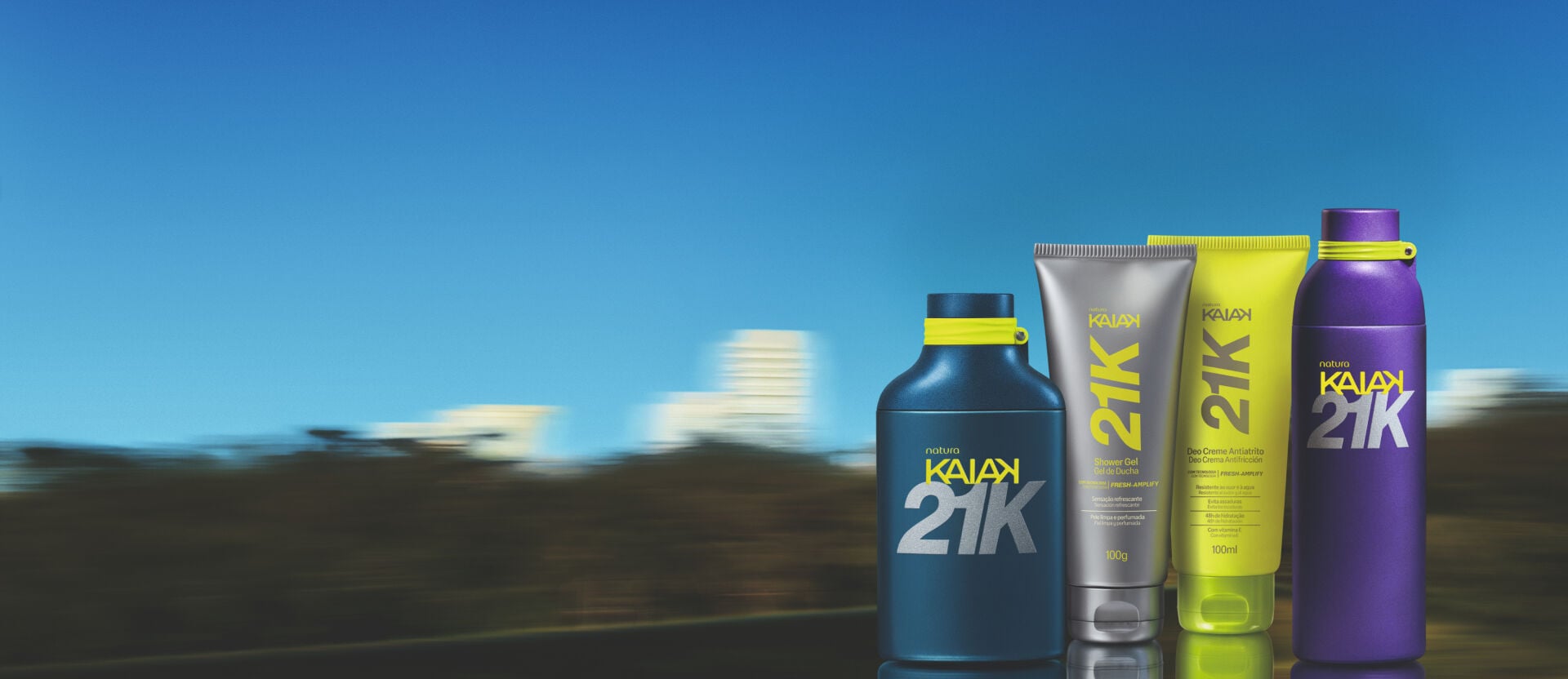 Imagem em fundo de paisagem desfocada com produtos Natura Kaiak 21K alinhados sobre reflexo. A linha esportiva traz perfume azul, shower gel cinza, protetor amarelo e garrafa roxa.