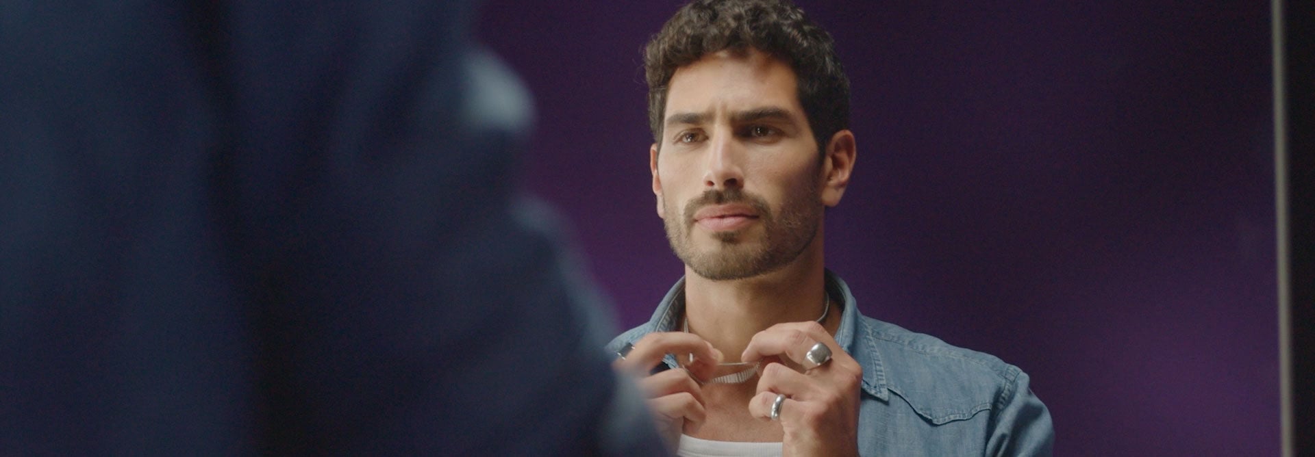 Homem branco de cabelo escuro e barba bem desenhada. Ele veste camisa jeans azul sobre uma camiseta branca e usa anéis, pulseira e colares prateados. Ele apresenta um frasco preto do Deo Parfum Natura Homem Identidad. Cenas do modelo arrumando as joias intercalam com momentos de interação com o produto, em que ele aparece borrifando, segurando e abrindo a fragrância.