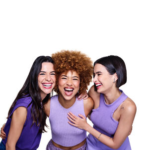 3 mulheres em fundo branco: uma negra de cabelo crespo ruivo, uma branca de cabelo escuro longo e uma branca de cabelo liso curto. Elas estão se abraçando, muito sorridentes, e todas vestem roxo.