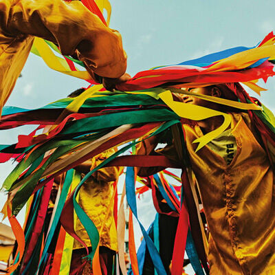 Vista em ângulo baixo de pessoas dançando em uma festividade cultural ao ar livre. Elas vestem camisas de cetim amarelo-ouro e calças vermelhas, segurando e agitando fitas coloridas e longas em tons de azul, verde, vermelho e amarelo. O céu ao fundo está claro com poucas nuvens, destacando o movimento dinâmico e alegre das fitas e dos dançarinos. 