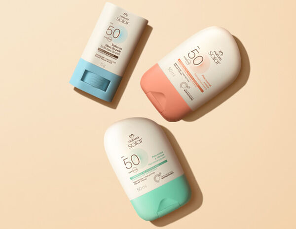 Três Protetores Solares Faciais FPS 50 Natura dispostos em diagonal. Todos têm embalagem branca, mas a base do stick é azul e as tampas dos demais são verde e laranja. Fundo em tom de creme.