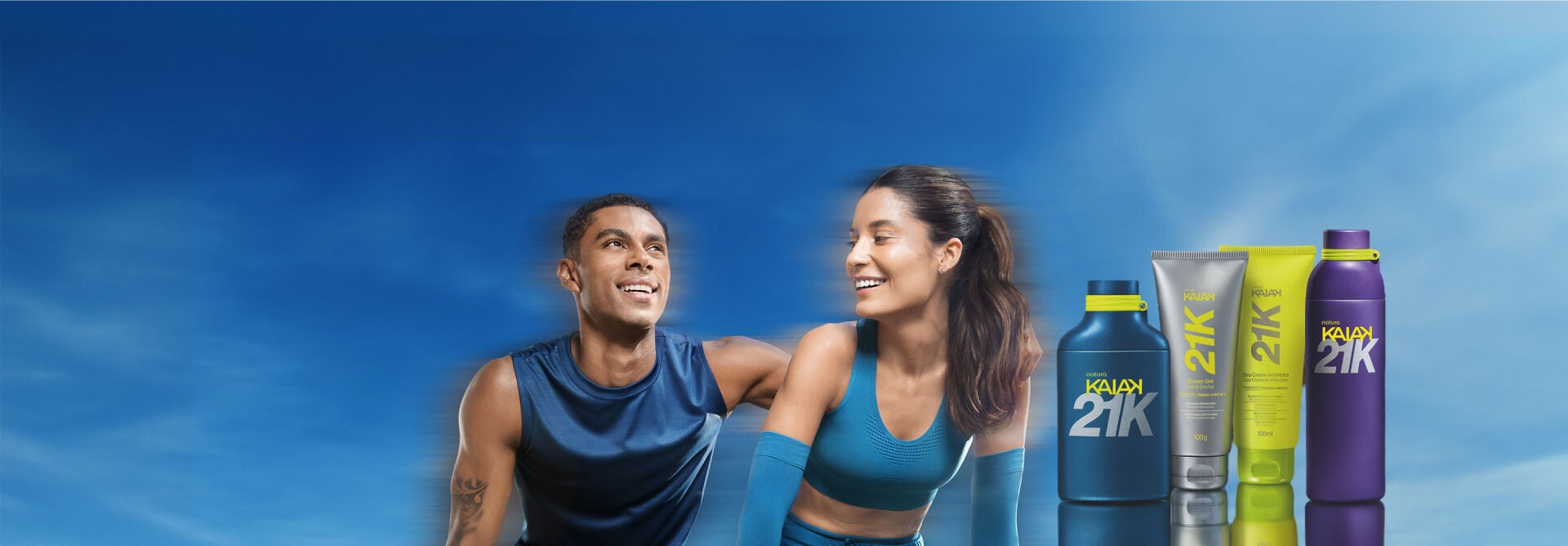 Banner sob céu azul com homem e mulher sorridentes em trajes de treino. À direita, frasco azul, shower gel, deo creme atrito e perfume roxo da linha Natura Kaiak 21K.