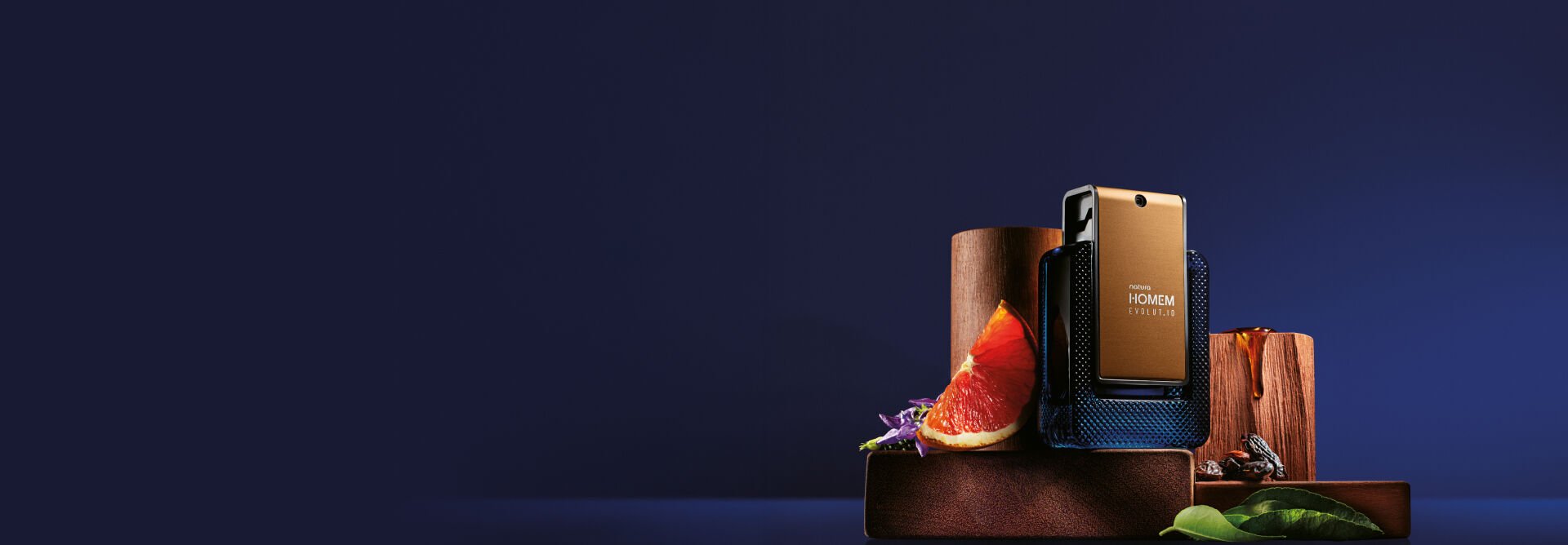 Frasco da fragrância Natura Homem Evolut.io sobre madeiras e cercado de frutas cítricas, flores, folhas e sementes. Fundo azul-escuro.