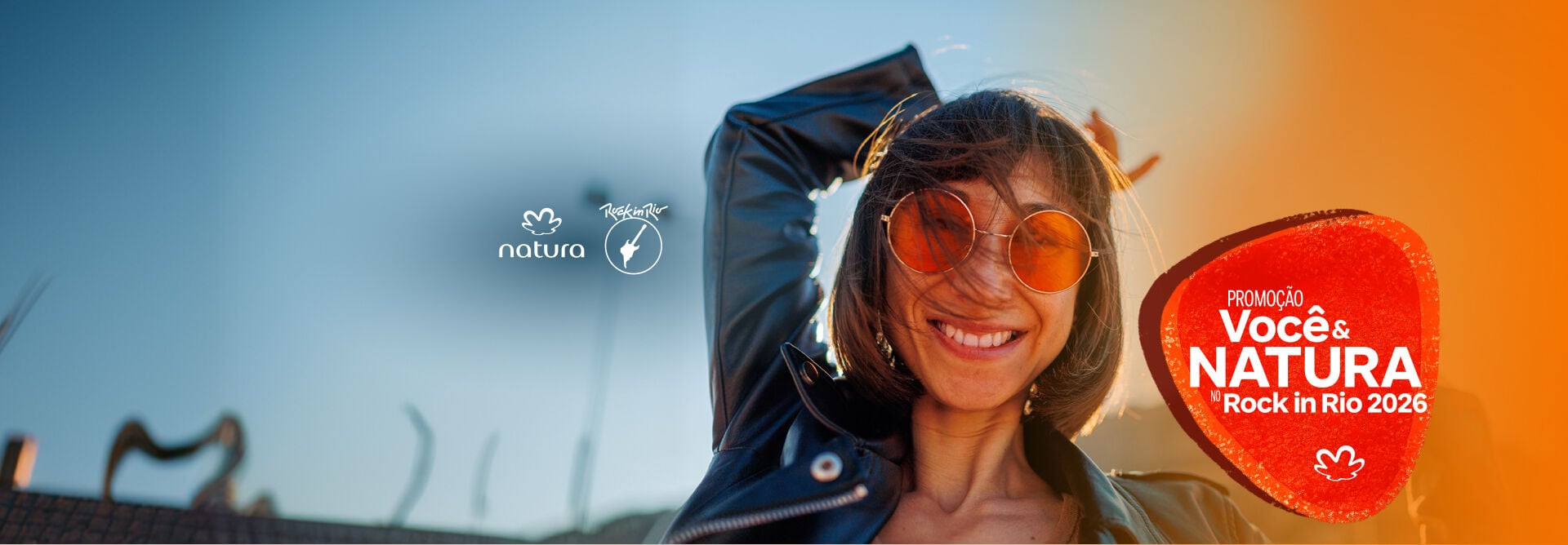 Mulher jovem sorridente vestindo jaqueta de couro e óculos escuros com lentes laranjas. Ao lado, o selo da promoção "Você & Natura no Rock in Rio 2026". 