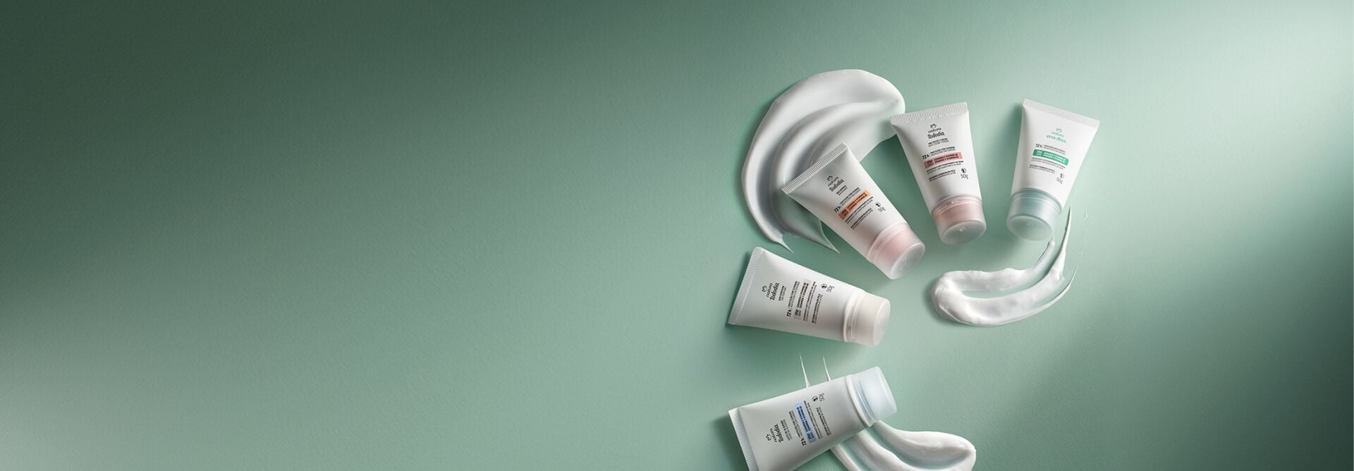 Quatro desodorantes em creme Natura Tododia e um Natura Erva Doce dispostos sobre superfície verde-água. As bisnagas brancas estão organizadas entre pinceladas de creme branco de textura macia. 