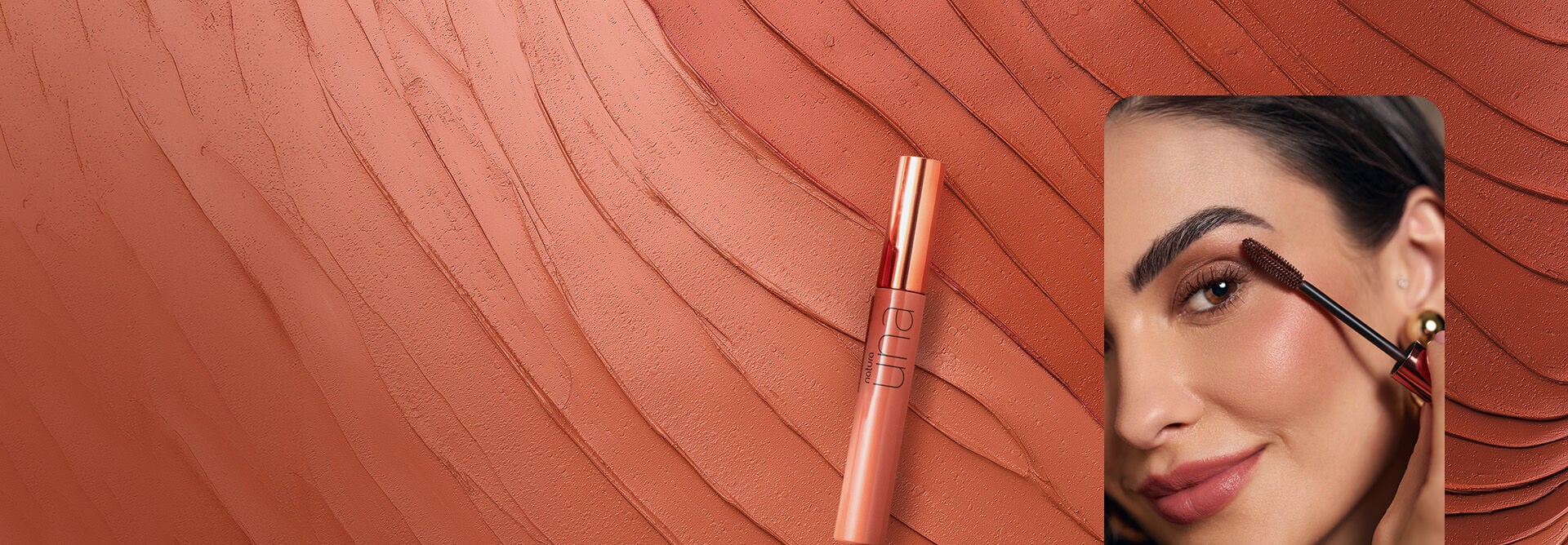 Tubo nude metalizado da Máscara para Cílios Volume Marrom Una, em um fundo com produto cremoso rosado espalhado em ondas. Ao lado, mulher branca de cabelo escuro aplica a máscara nos cílios.