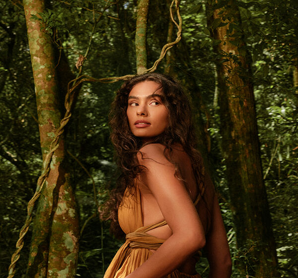 Mulher em uma floresta cercada por árvores altas. Usa vestido marrom, tem cabelos castanhos longos e cacheados e maquiagem em tons naturais, com pele iluminada e lábios nude.