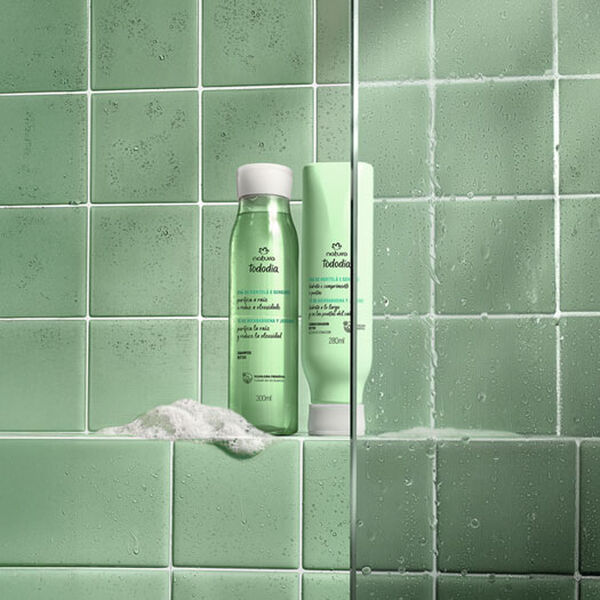 Frascos verdes com tampas brancas de Shampoo e Condicionador da linha Tododia Chá de Hortelã e Gengibre. Eles estão sobre uma espuma branca, em um banheiro com azulejos verdes e box de vidro.