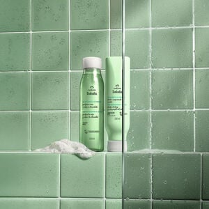 Frascos verdes com tampas brancas de Shampoo e Condicionador da linha Tododia Chá de Hortelã e Gengibre. Eles estão sobre uma espuma branca, em um banheiro com azulejos verdes e box de vidro.