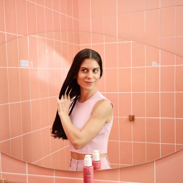 Mulher de pele clara e regata rosa espalha leave-in pelos fios lisos e úmidos em frente ao espelho. Ao fundo, azulejos rosa e, em destaque no balcão, embalagem da linha Natura Tododia Liso + Liso. 