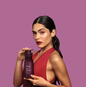 Mulher de cabelos castanhos e presos usando vestido vermelho segura um produto Natura Tododia, em fundo lilás.