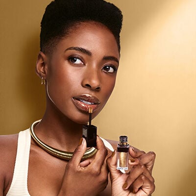 Mulher negra de cabelo crespo curto em fundo dourado, usando regata branca e um colar dourado. Ela segura um frasco dourado com aplicador marrom e deposita gloss labial dourado nos lábios.