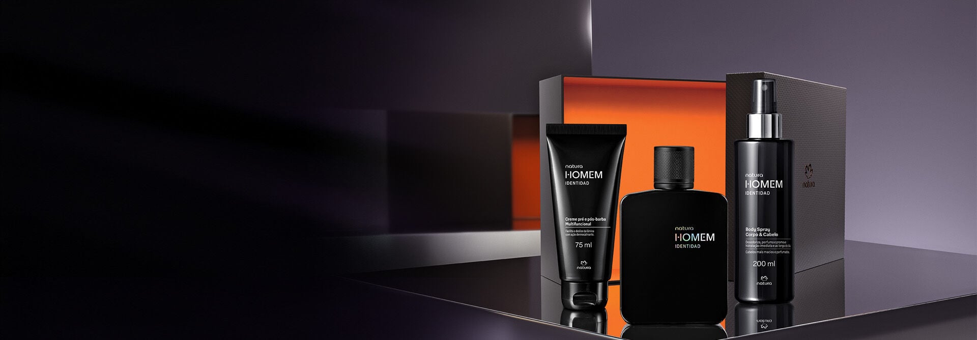 Produtos Natura Homem Identidad: Creme Pré e Pós-Barba Multifuncional, Deo Parfum e um Body Spray Corpo & Cabelo. Embalagens pretas junto de uma caixa laranja e cinza, sobre plataforma preta reflexiva.