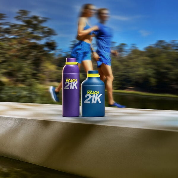 Perfume Natura Kaiak 21K em tons de roxo e azul repousam em mureta. Ao fundo desfocado, duas pessoas correm ao ar livre, transmitindo movimento, energia e desempenho esportivo.
