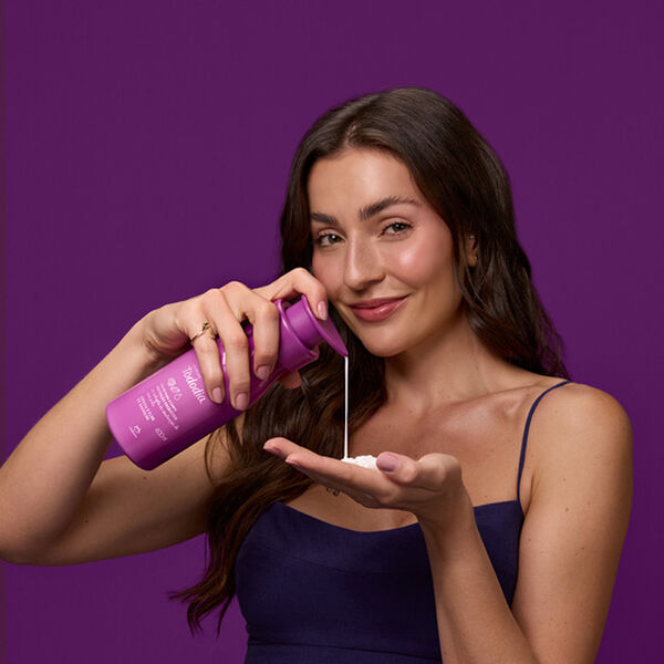 Mulher branca de cabelo castanho segura um frasco roxo com tampa pump do Creme Desodorante Nutritivo para o Corpo Tododia Ameixa e Flor de Baunilha. Em fundo roxo, ela despeja creme branco na mão.
