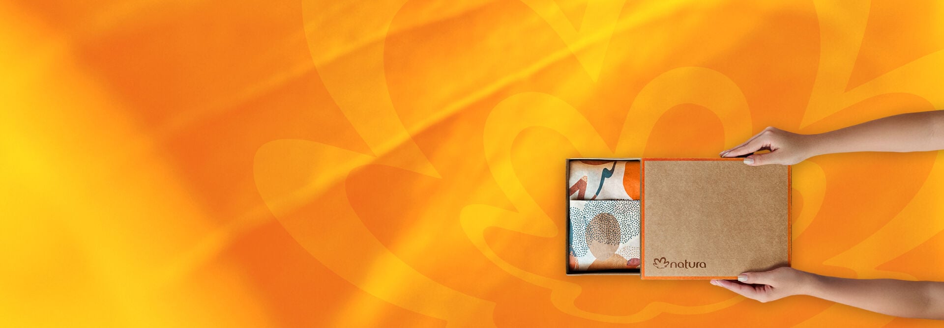 Na imagem, mãos femininas abrem um presente Natura, com caixa parda, logo Natura, detalhes em laranja e papel de presente decorado. Fundo em tons de amarelo e laranja com a flor-símbolo da Natura.