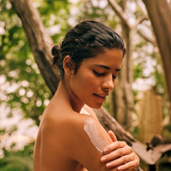 Mulher morena com cabelos presos aplica Polpa Desodorante Hidratante para o Corpo Ekos Cacau nos ombros em meio à floresta.