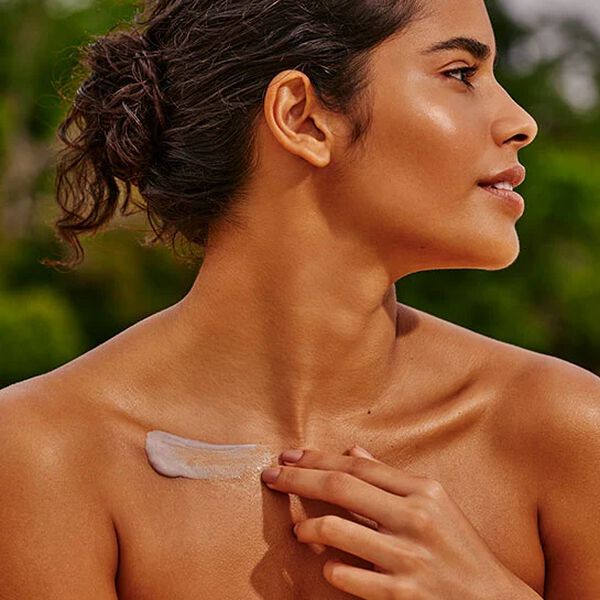 Mulher de pele morena e cabelo castanho preso aplica creme branco na região da clavícula. Ela está ao ar livre, com fundo desfocado de vegetação verde.