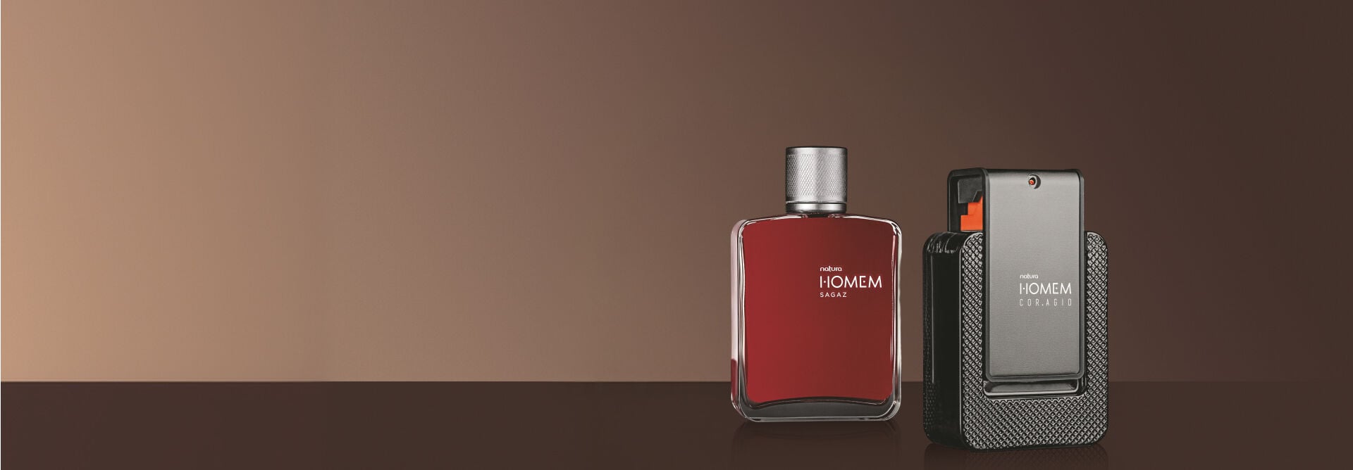 Dois frascos de Natura Homem estão sobre superfície fundo marrom. À esquerda, o Homem Sagaz em frasco vermelho-fechado; à direita, o Homem Cor.agio em embalagem cinza-chumbo e preta.
