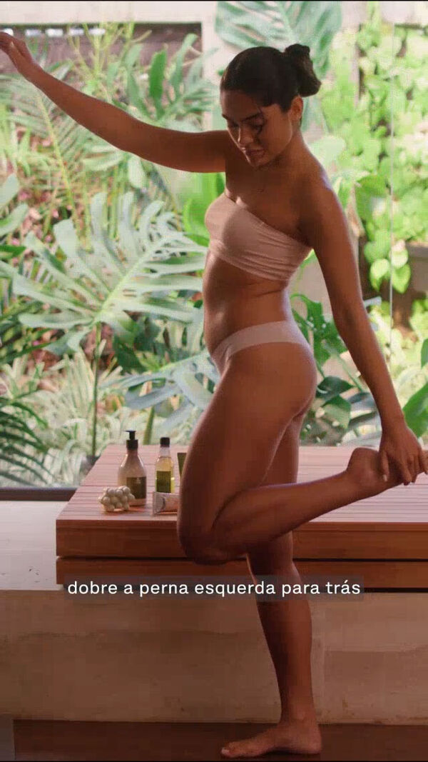 Em cenário de jardim tropical, vídeo de produtos da linha Ekos Andiroba em bancada em madeira, com mulher de cabelos castanhos e presos, usando biquíni branco, fazendo alongamento corporal. Ela também faz massagem nas coxas, pernas e pés. Finaliza com mulher respirando fundo e destaque para itens da linha.