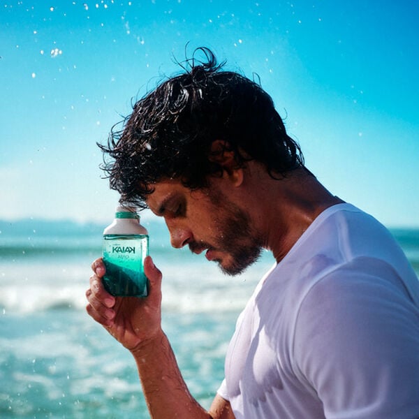 Homem de cabelos escuros e camiseta branca na praia com mar e céu azul ao fundo. Ele segura junto à têmpora frasco verde com gotas de água na embalagem da fragrância Kaiak Aero.
