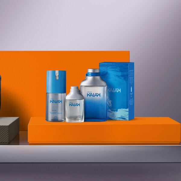 Linha de perfumaria masculina Natura Kaiak disposta sobre um bloco laranja, composta por desodorante spray, miniatura de colônia, o perfume clássico em frasco azul e prata, e sua caixa azul ao fundo.
